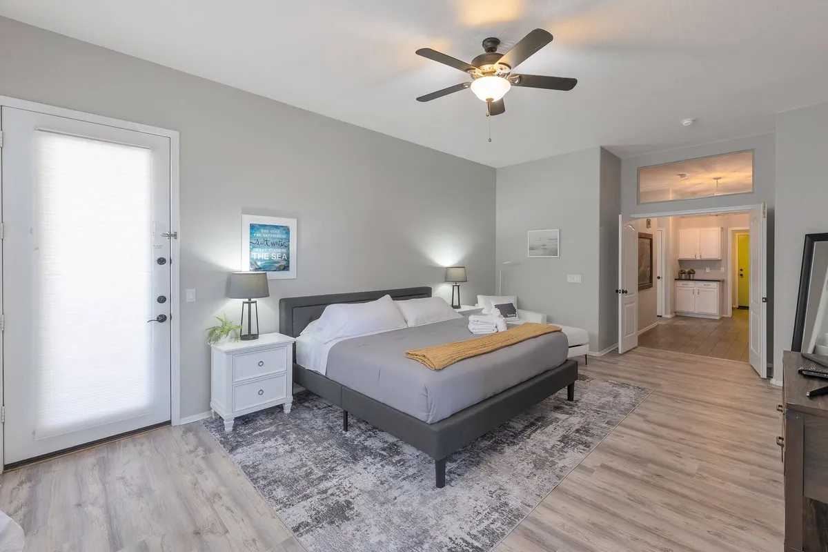 A tranquil bedroom with a bed dressed in gray bedding, matching nightstands with lamps, artwork above the bed, and a door leading outside. The space is filled with natural light, offering a calm and restful atmosphere.