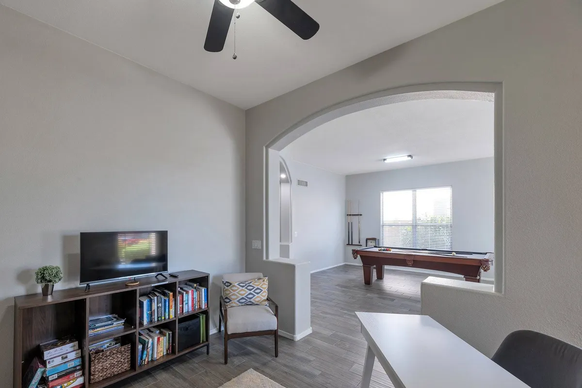 A casual and inviting entertainment room featuring a pool table, large windows streaming in natural light, a television, bookshelf, and a desk. The space is versatile—ideal for memorable game nights, movie marathons, or a quiet afternoon with a book.