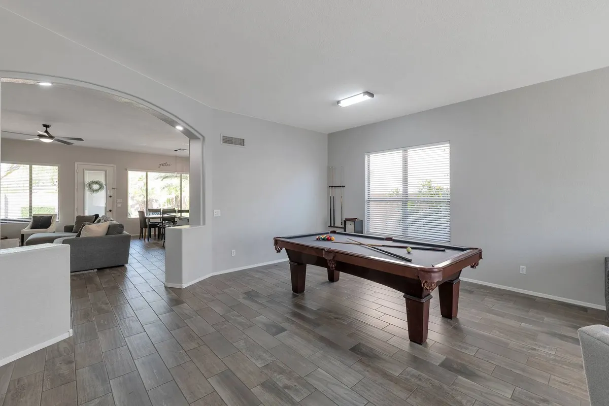 This image presents a welcoming game room featuring a pool table at the center, surrounded by comfortable sofas and bathed in natural light from large windows. The arched doorway adds architectural interest, and the arrangement clearly encourages both socializing and play.