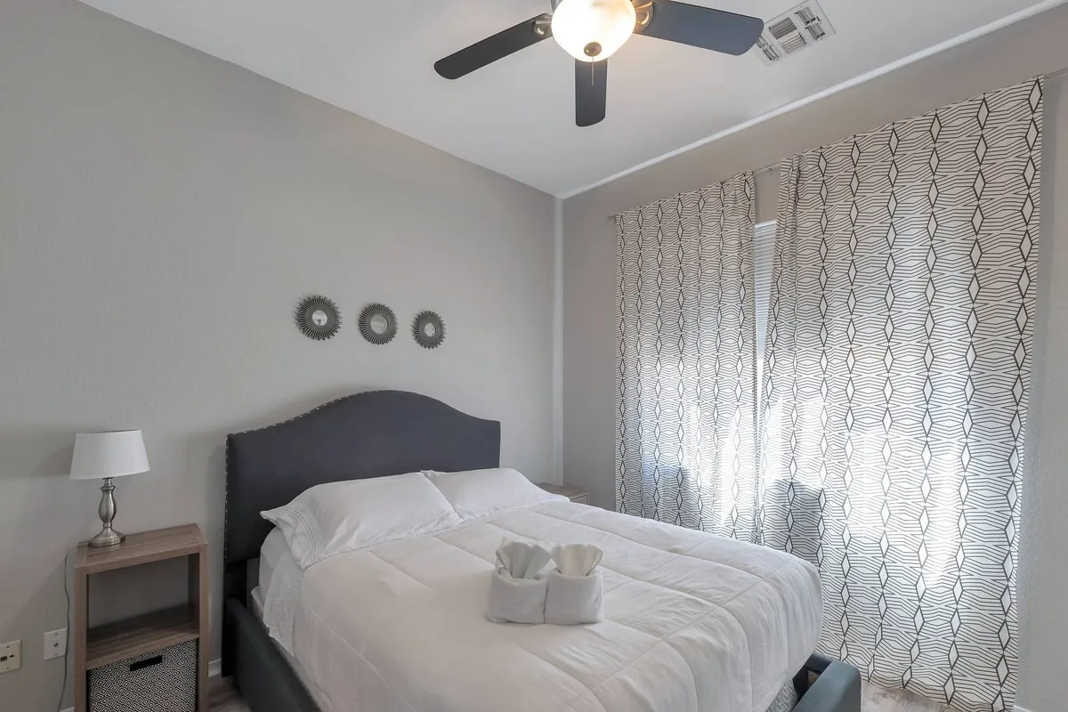 A serene bedroom with a bed dressed in crisp white linens, decorative mirrors on the wall, geometric-patterned curtains, and a nightstand with a lamp. The space is filled with soft natural light, creating a tranquil and comfortable retreat for rest and relaxation.