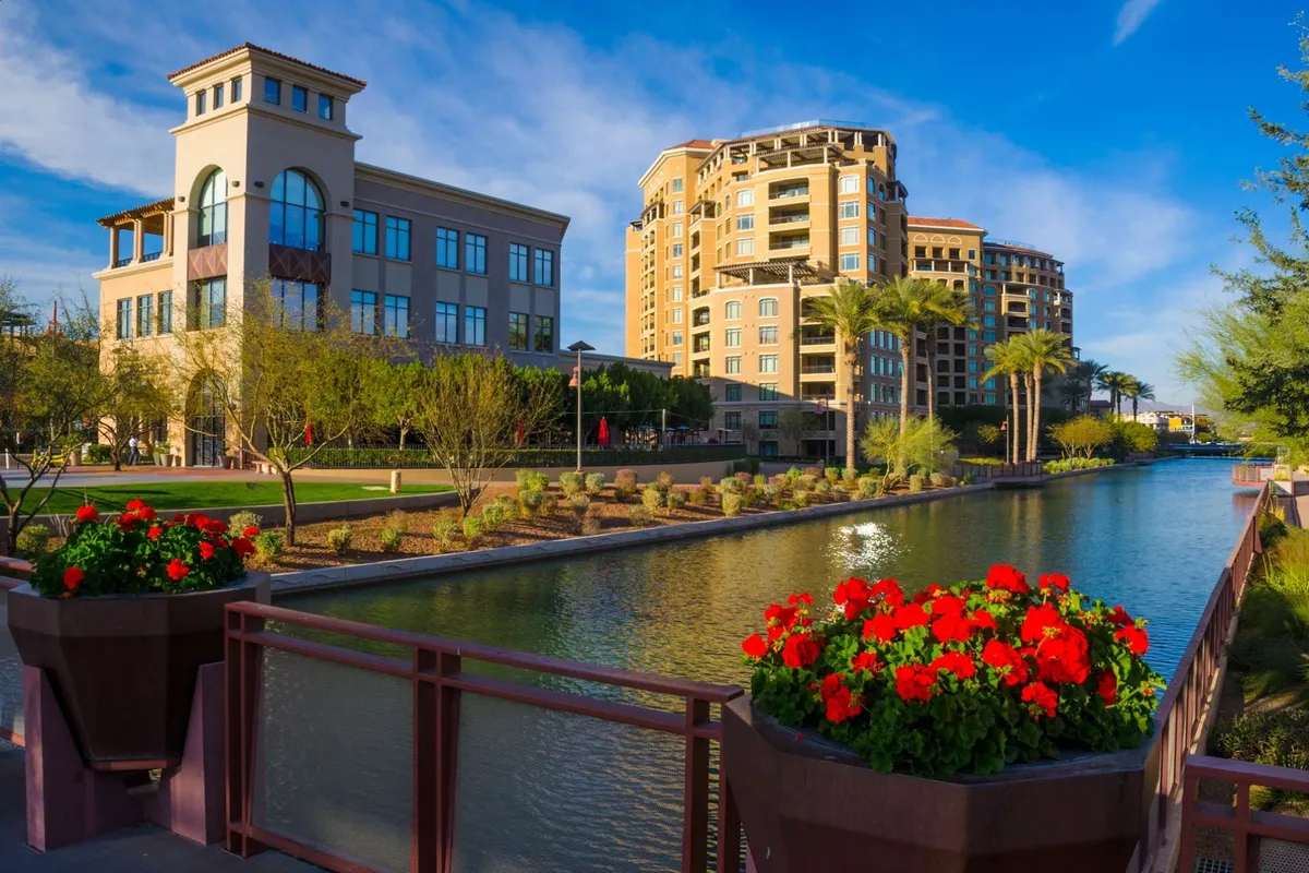 A vibrant exterior scene with modern architecture, a scenic canal, flower planters, and palm trees. People can be seen strolling along the canal and enjoying outdoor dining, emphasizing the lively neighborhood atmosphere and proximity to attractions.