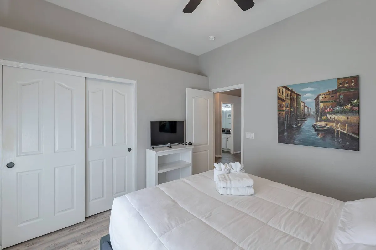 A guest bedroom with a comfortable bed adorned in white linens, vibrant artwork on the wall, and a TV with a modern stand. The mood is calm and cozy, making it an inviting space for relaxation and enjoying a leisurely breakfast or movie night.