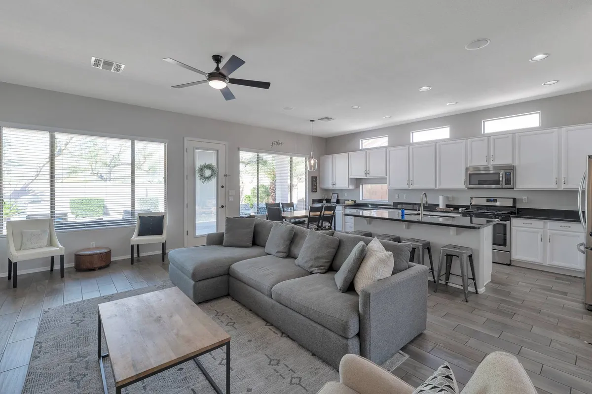 The image showcases a bright, open-concept living space featuring a spacious grey sectional perfect for groups, a modern kitchen island, and a cozy dining area, all bathed in sunlight streaming through expansive windows.