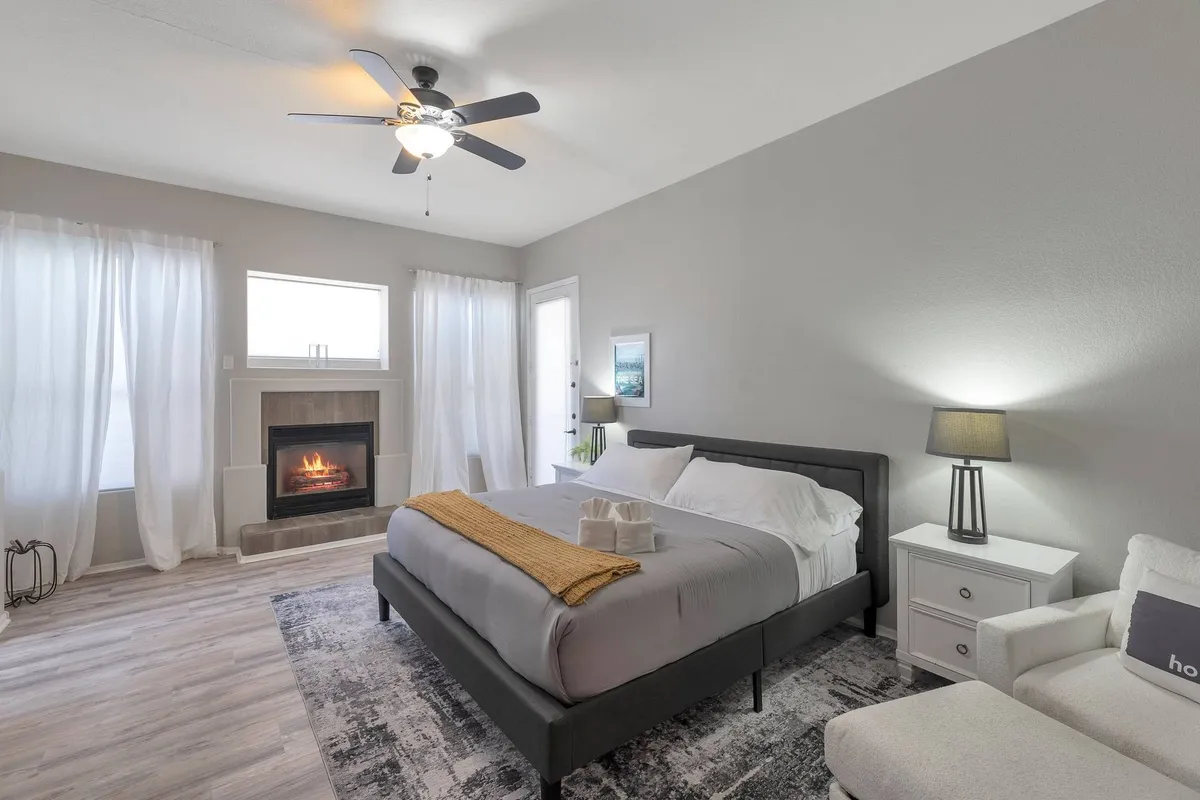 A spacious master bedroom featuring a king bed with gray linens and a vibrant yellow throw blanket. The room includes a cozy fireplace, large windows draped with sheer curtains letting in natural light, and a stylish nightstand with a lamp. The atmosphere is tranquil and inviting—ideal for relaxing by the fireplace or enjoying a morning coffee in bed.