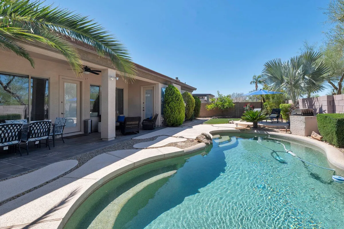This image features a sparkling swimming pool bordered by lush greenery and palm trees, with a patio seating area nearby. The scene is infused with bright sunlight, highlighting the pool and inviting relaxation in the lounge chairs or patio space.