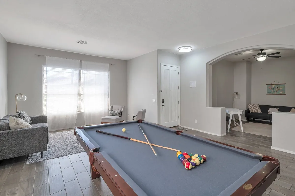 An interior shot of a bright, casual game room featuring a pool table, windows streaming in natural light, a sofa, and the entrance door. The space is set up for friendly competition, socializing, and family fun.