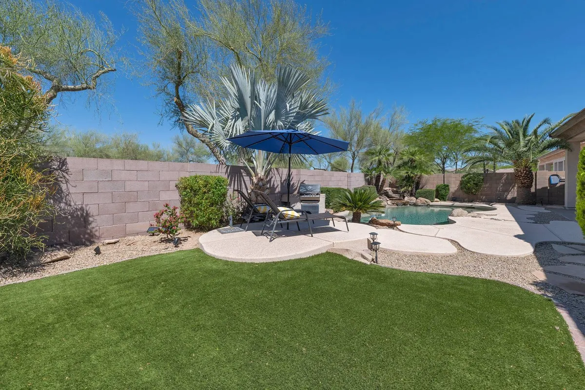 A tranquil pool scene with an umbrella providing shade over lounge chairs, polished landscaping surrounding the water, and hints of outdoor dining possibilities. The mood is serene, suggesting both sun-soaked days and relaxing evenings by the pool.