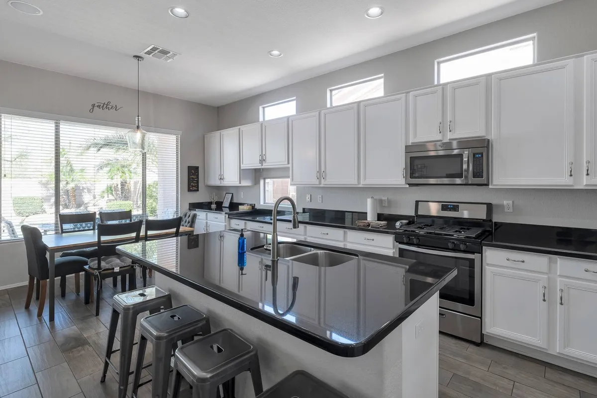 This image captures a bright, inviting kitchen with a central island countertop, stools, visible kitchen appliances, and windows letting in natural light. The scene conveys activity, with ample space for cooking and socializing, and highlights the breakfast bar seating.